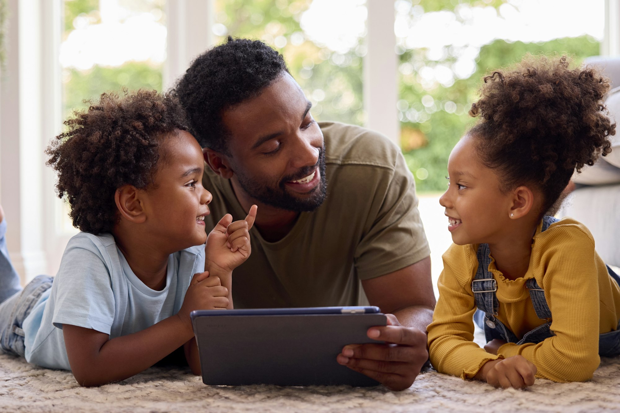 Father At Home Lying On Rug In Lounge With Children Using Digital Tablet